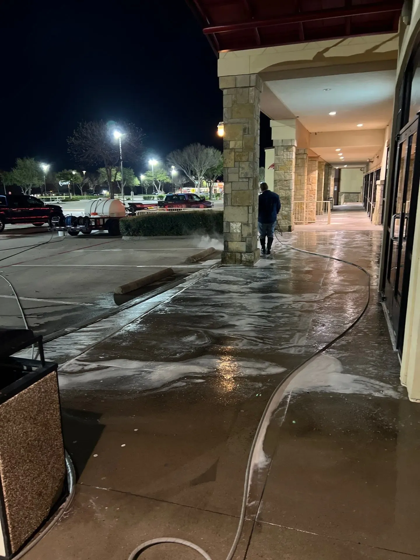 Technician rinsing a shopping center sidewalk during an overnight pressure washing job with the wash rig staged nearby in Frisco, TX